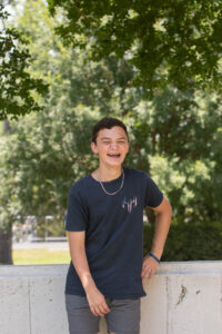 A teenage boy with short brown hair smiles widely, showing his braces, as he stands outside on a sunny day. He is wearing a dark T-shirt with a small American flag design and gray shorts. The background features lush green trees, reflecting the positive impact RMHC has in supporting families during medical crises and helping them find moments of joy.