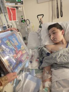 A teenage boy lies in a hospital bed, surrounded by medical equipment and tubes, including a nasal tube. He is looking at a clear plastic container filled with various snacks being held by a person out of frame. The boy is recovering in a hospital setting, with a heart monitor and other medical devices visible in the background, symbolizing how RMHC supports families during medical crises with care and essentials.