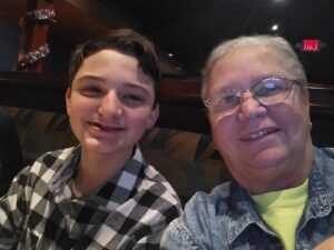 A smiling teenage boy with short brown hair, wearing a black and white checkered shirt, sits next to an older man with glasses and short gray hair, who is also smiling. The two are enjoying a moment together in a cozy, warmly lit movie theater, reflecting how RMHC is supporting families during medical crises by providing comfort and care.