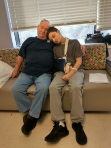 An older man and a teenage boy sit closely together on a couch, both with their eyes closed. The boy, wearing a gray shirt and sweatpants with one arm in a sling, rests his head on the man’s shoulder. The man, dressed in a navy blue shirt and jeans, has his arm around the boy, providing comfort, illustrating the support RMHC provides to families during medical crises by offering a space for rest and recovery.